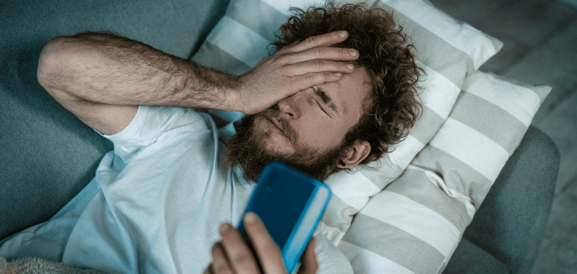 Man holding phone, lying on couch, looking tired.